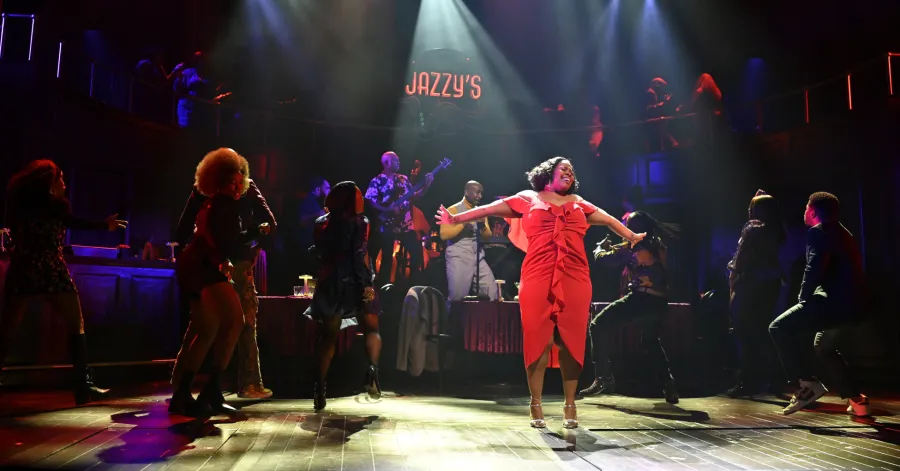 Performers dance energetically on stage at a jazz club set, with a singer in a red dress in the spotlight and "Jazzy's" sign illuminated above.