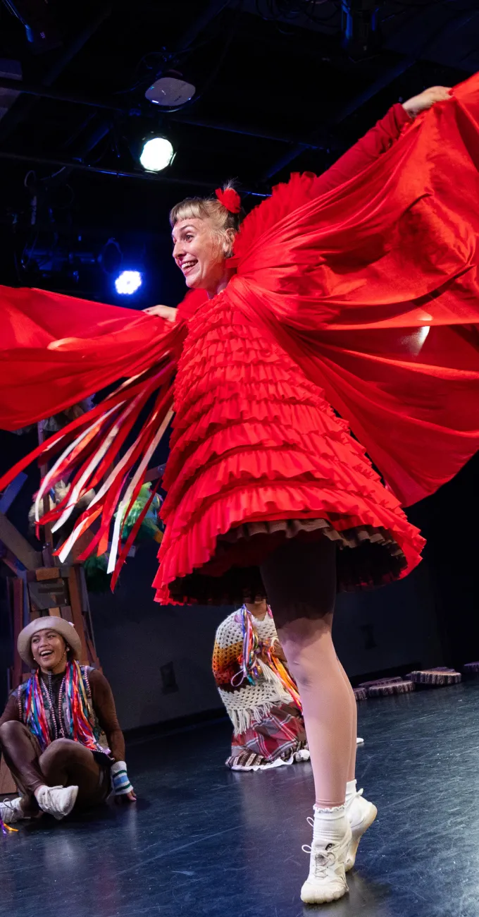 Performer in a bright red, ruffled costume with outstretched sleeves resembling wings, smiling on stage with two seated performers in the background.