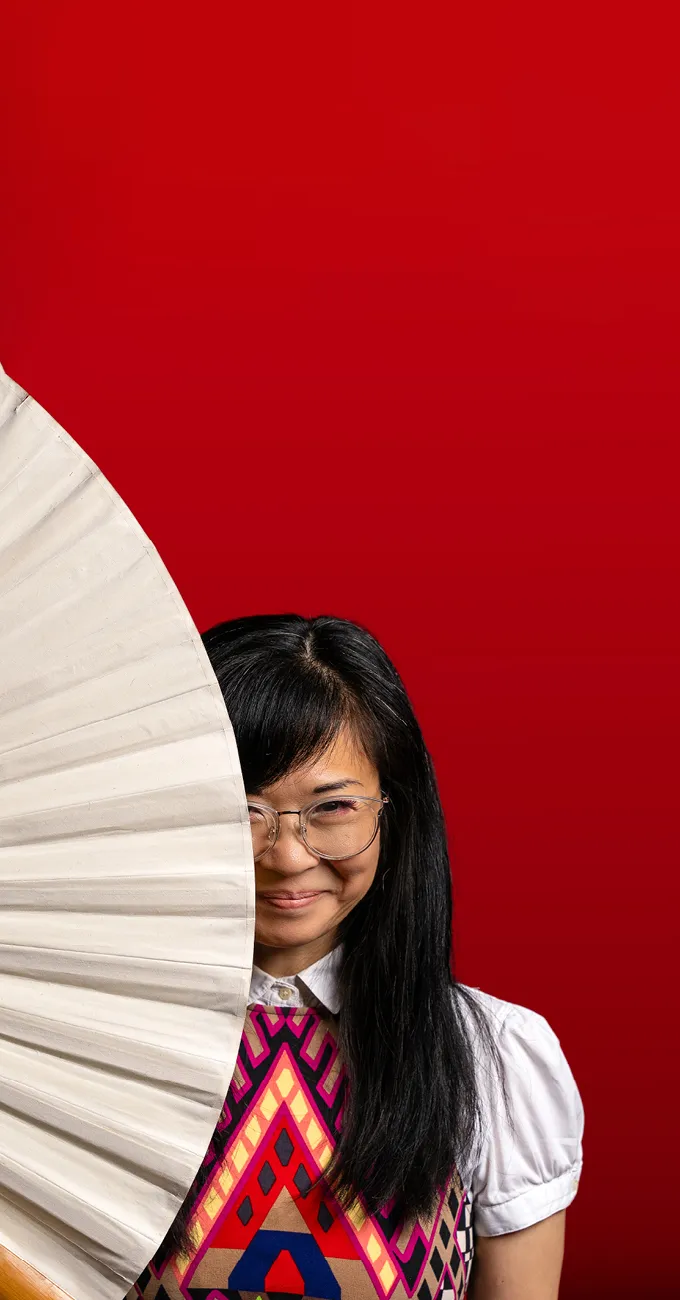 A woman with long black hair and glasses stands against a red background, holding a large white fan that covers half of her face.