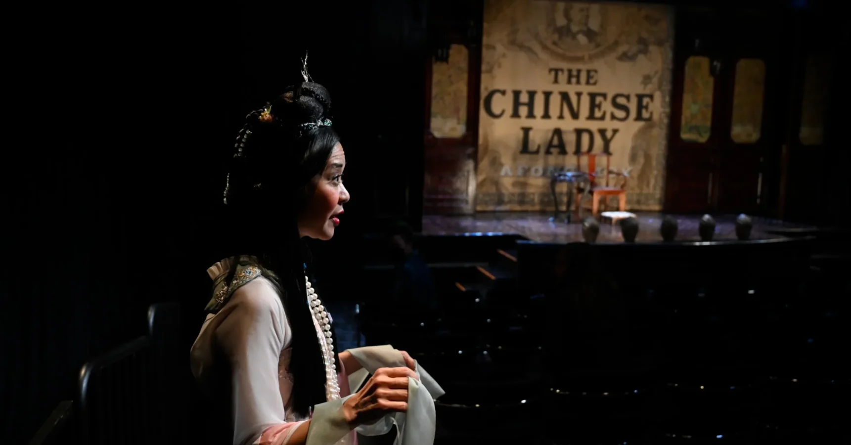 A woman in traditional Chinese attire stands in a darkened theater, facing a stage with a backdrop that reads "The Chinese Lady.
