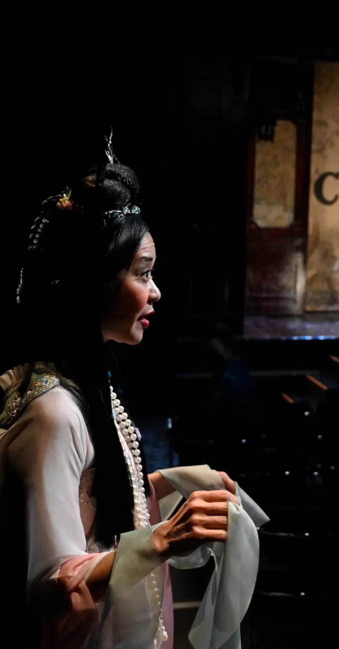 A woman in traditional Asian costume stands on stage, holding fabric, with dim lighting and a partially visible audience in the background.