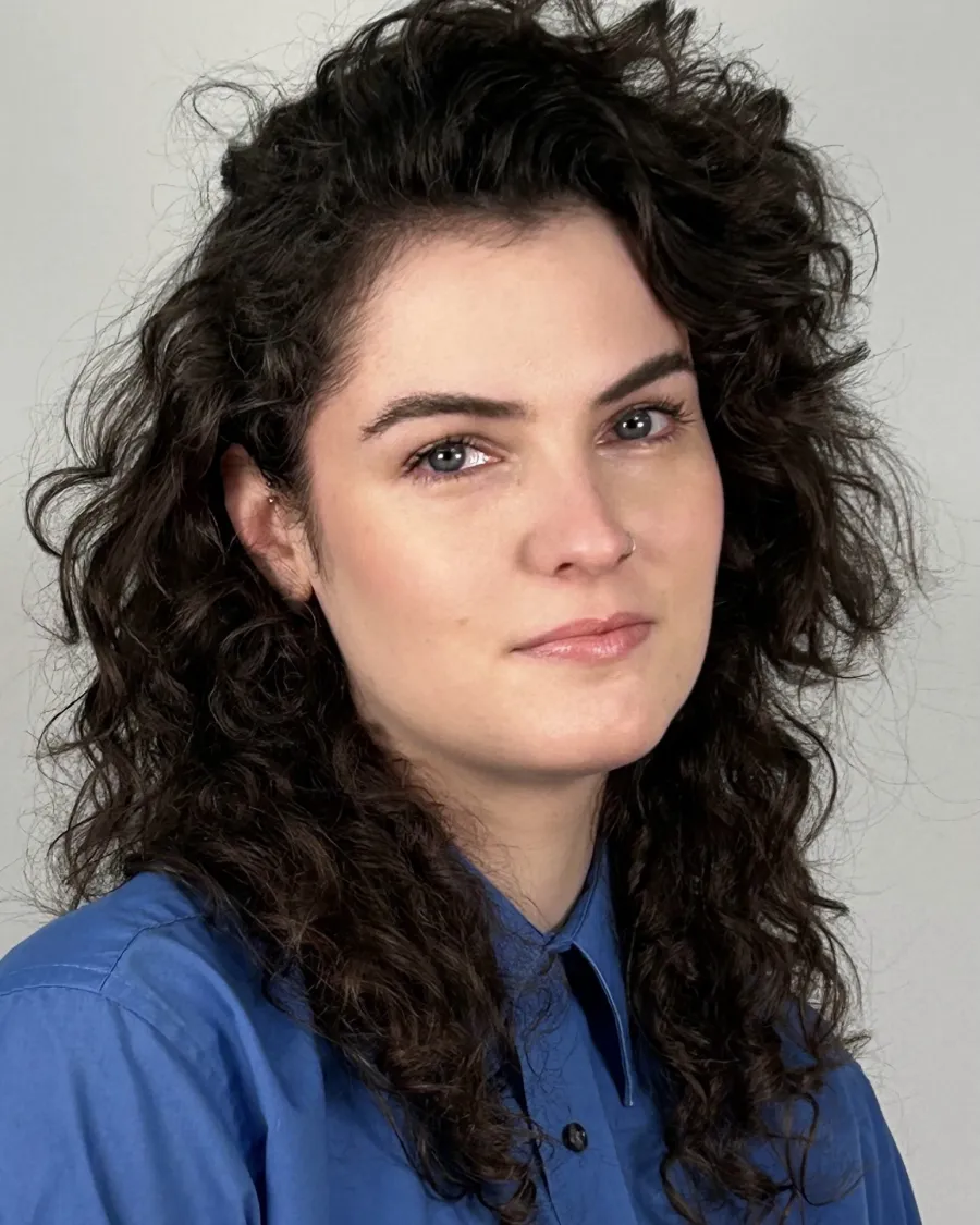 A person with long, curly dark hair and light skin wears a blue collared shirt and looks at the camera against a plain light background.