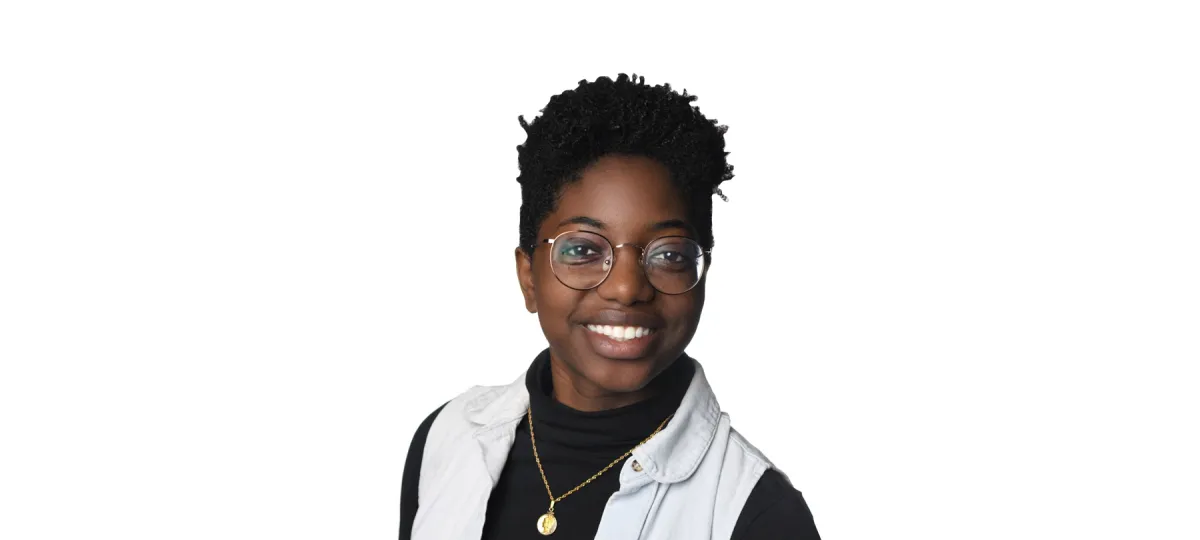 A person with short curly hair, glasses, and a black turtleneck layered with a light vest, smiling against a plain white background.