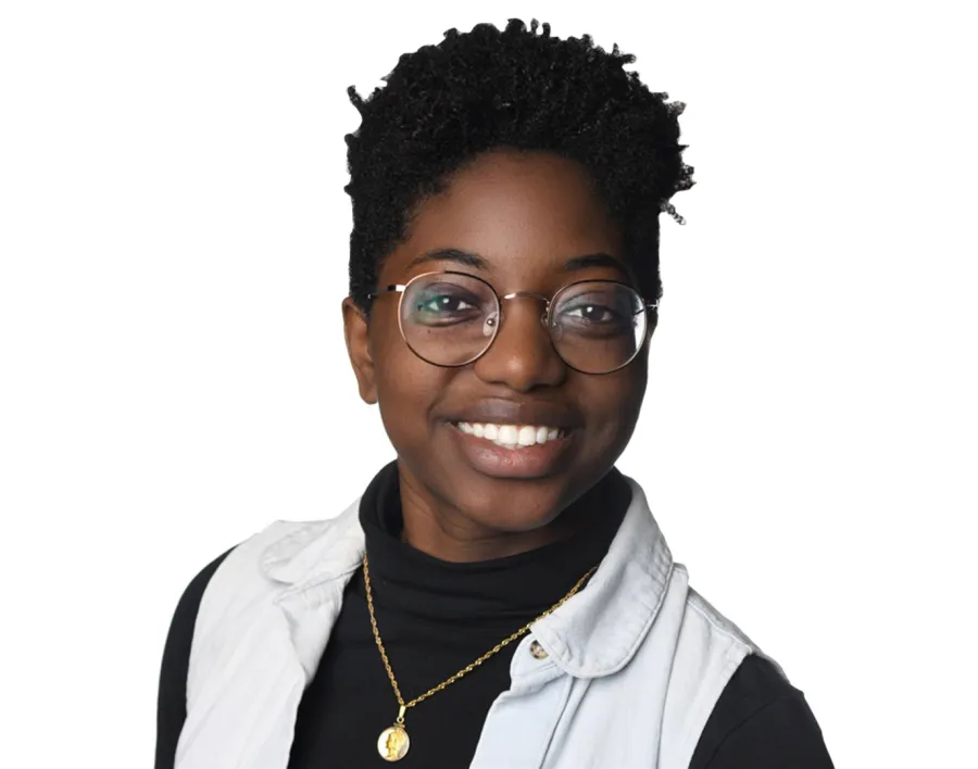 A person with short curly hair, glasses, and a black turtleneck under a light denim vest, smiling at the camera against a white background.