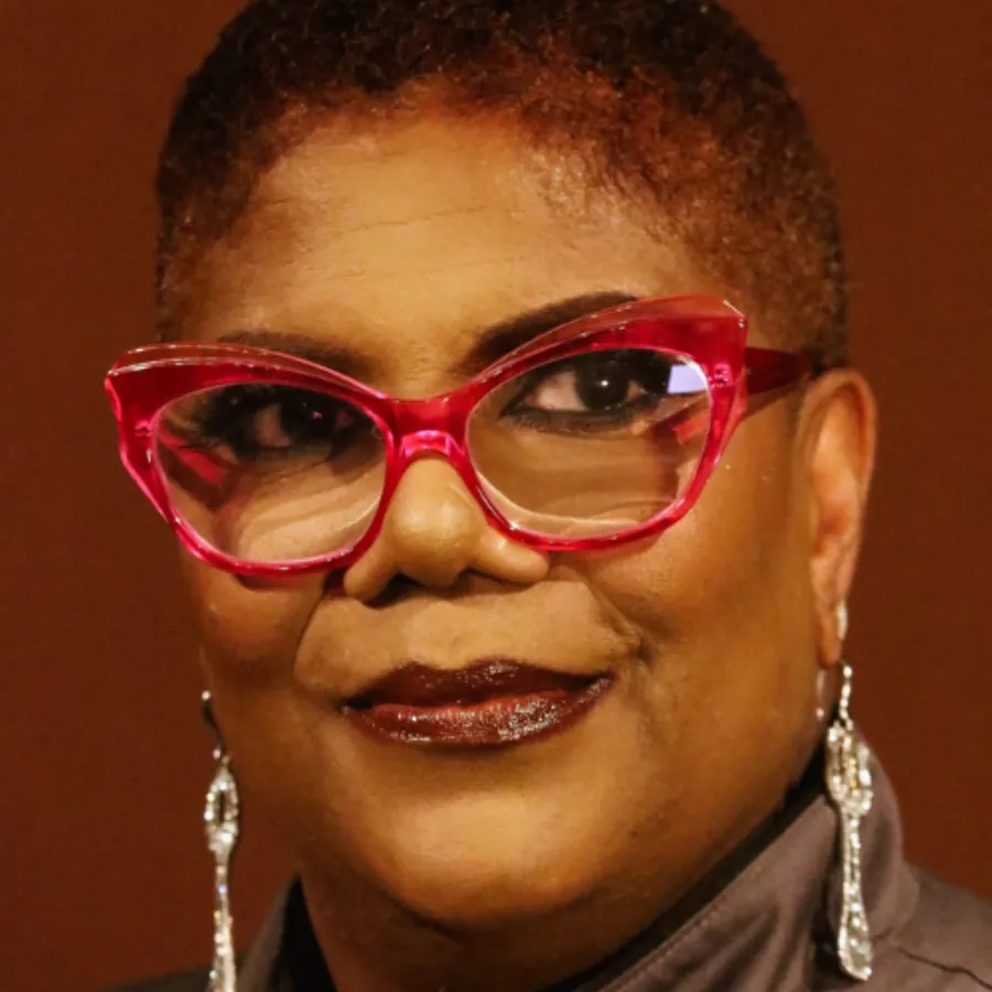 A person with short hair, wearing bold red glasses, long dangling earrings, and dark lipstick, looks toward the camera against a brown background.