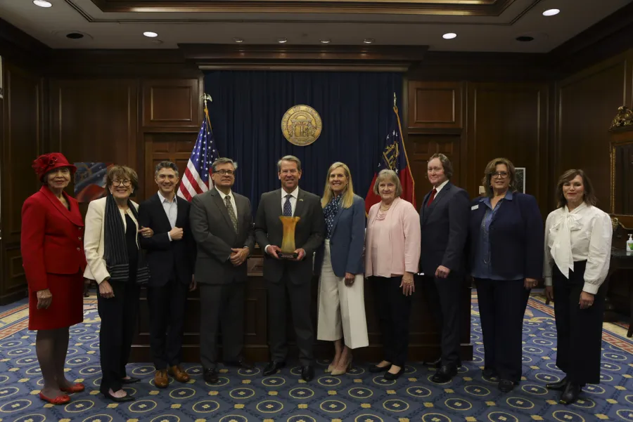 A group of ten people stands in a formal room with flags and a seal behind them; the man in the center holds a wooden award.