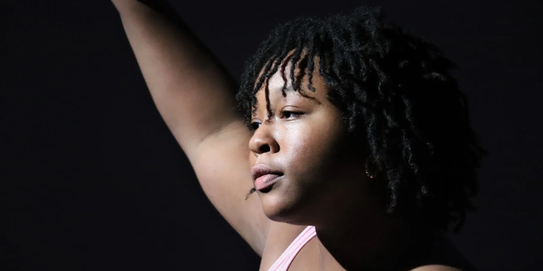 A person with short, curly hair raises one arm, looking thoughtfully into the distance against a dark background.
