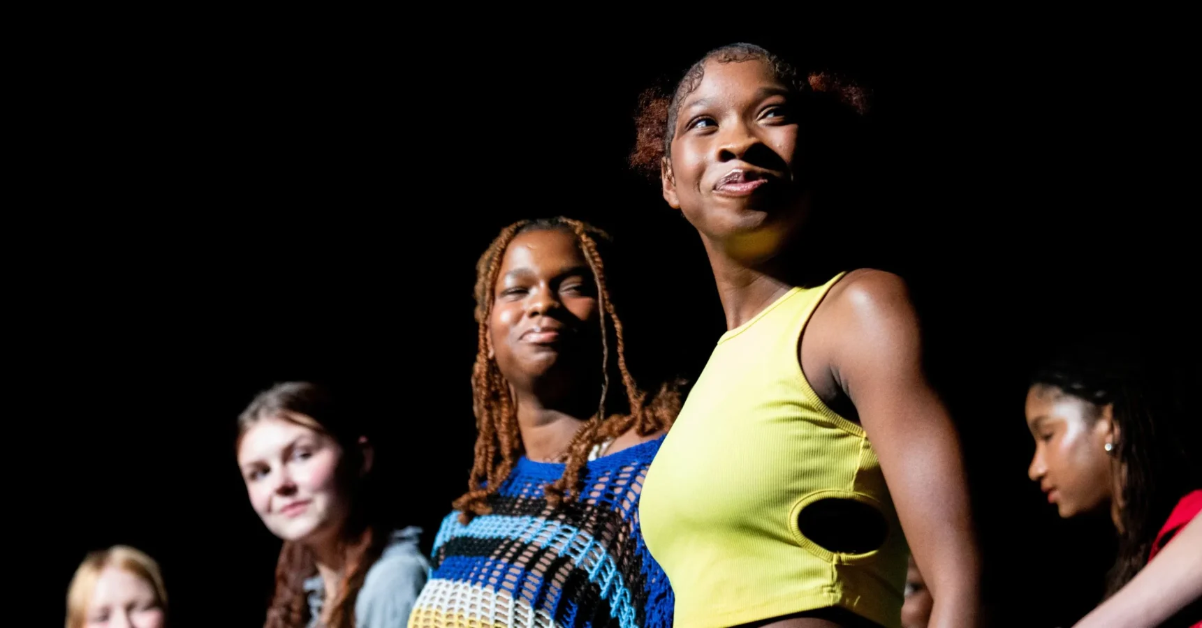 A group of young women stand together onstage, facing forward under bright lighting against a dark background.