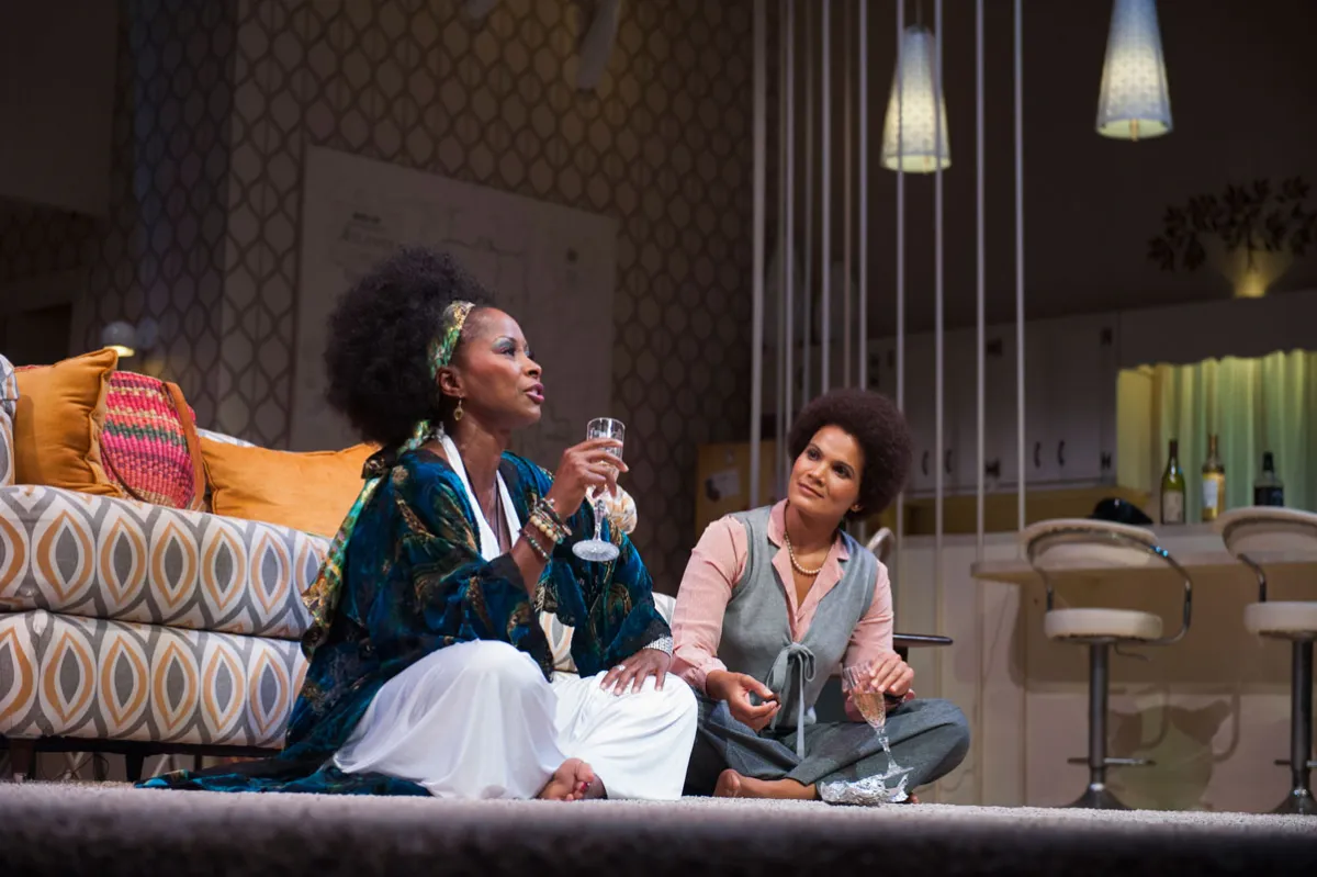 Two women sit on a carpeted floor in a stylish living room, talking and drinking from wine glasses; one is dressed in a robe, the other in casual attire.