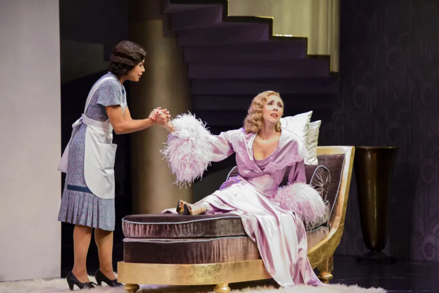 A woman in a maid uniform holds hands with a woman in an elegant robe who is seated on a chaise lounge, appearing distressed, in a dramatic stage setting.