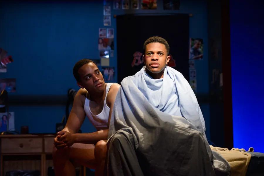 Two young men sit on a bed; one is wrapped in a sheet and looks ahead, while the other sits beside him in a tank top, looking at him. The room has blue walls and photos on the wall.