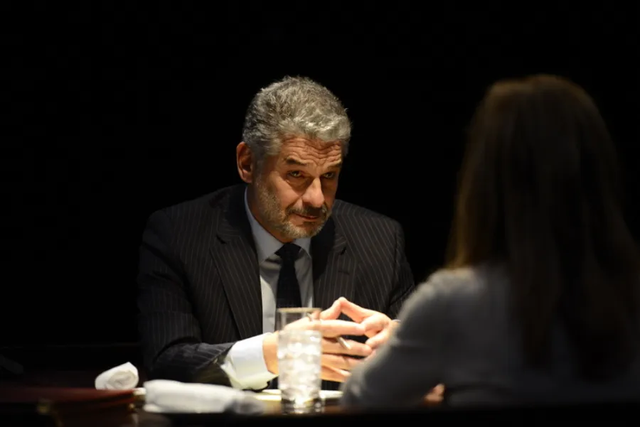 A man in a suit sits at a table across from a woman, with his hands clasped and a glass of water in front of him, under focused lighting.