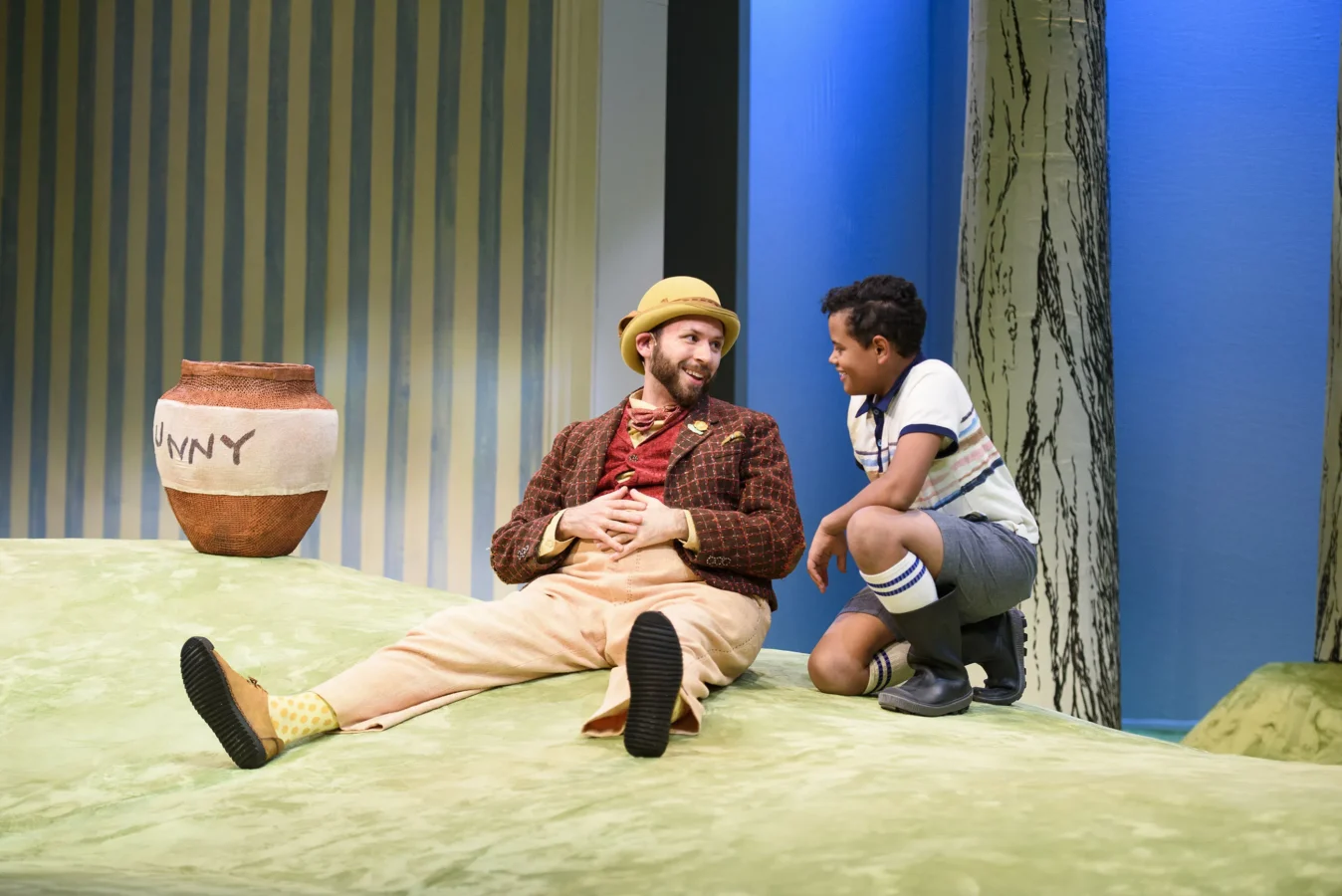 An adult in a checked jacket and yellow hat sits on a grassy stage set, smiling at a child in shorts and suspenders; a large honey pot is nearby.