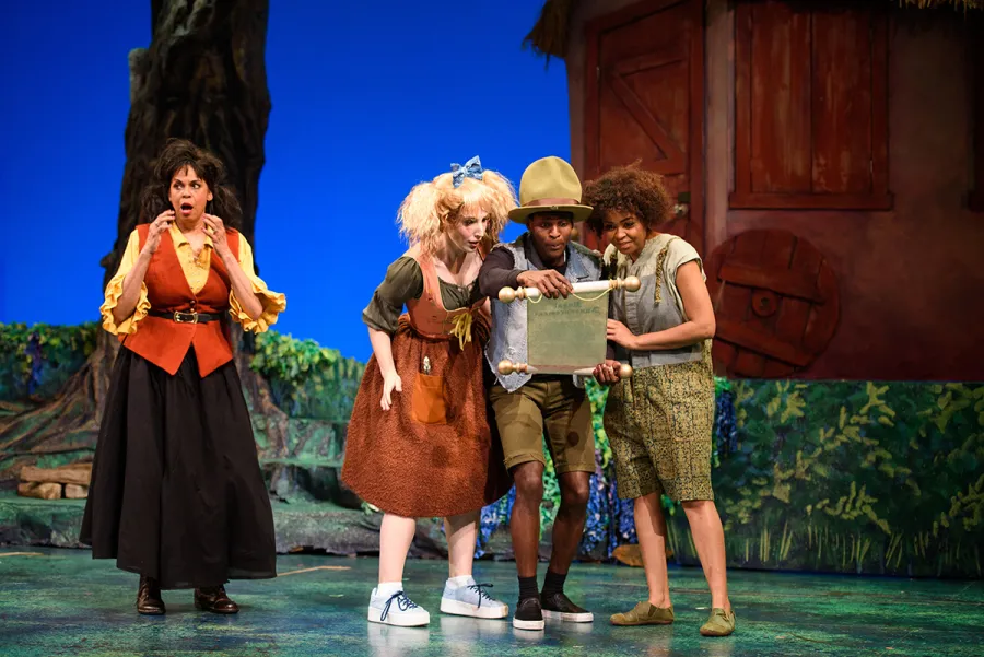 Four actors in colorful costumes perform on stage; two hold a scroll while the others react, with painted trees and a wooden house backdrop behind them.