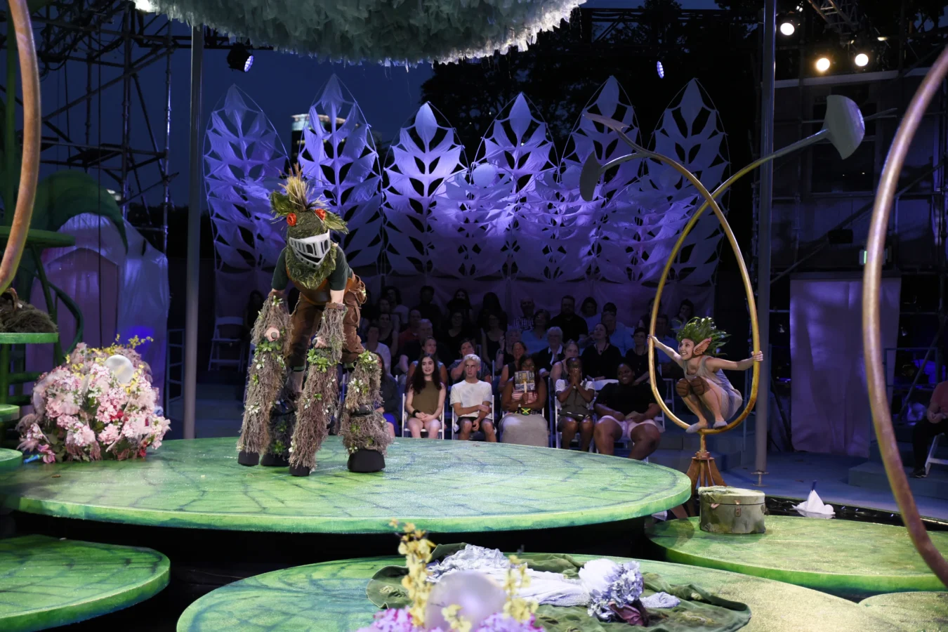 Actors in elaborate costumes perform on a stage with green platforms and floral decorations before an audience; one actor is on stilts, another is seated on a suspended ring.