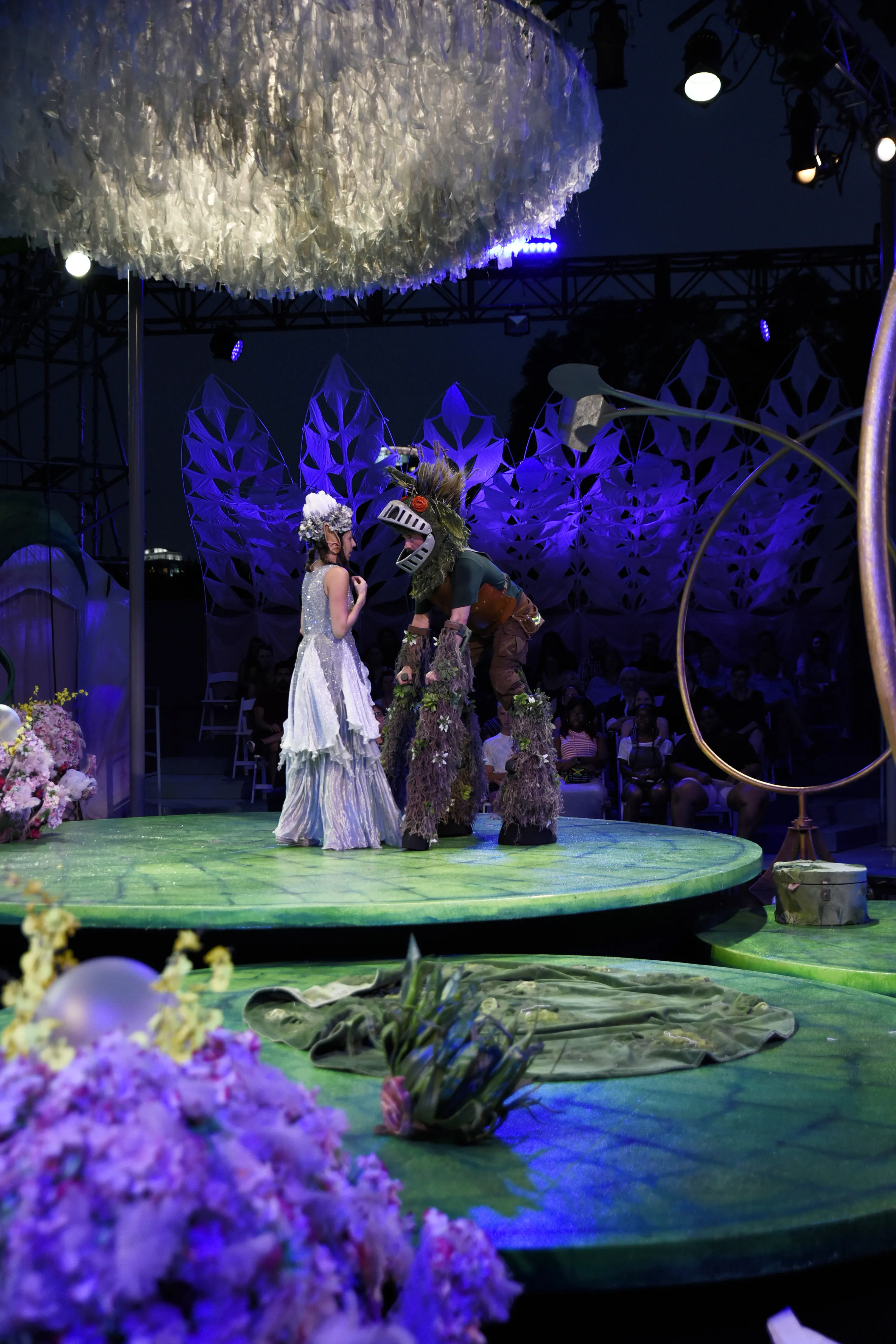 Two actors in elaborate costumes, one wearing a white dress and the other in a mossy, horned outfit, perform on a green-lit stage with floral decorations and dramatic lighting.