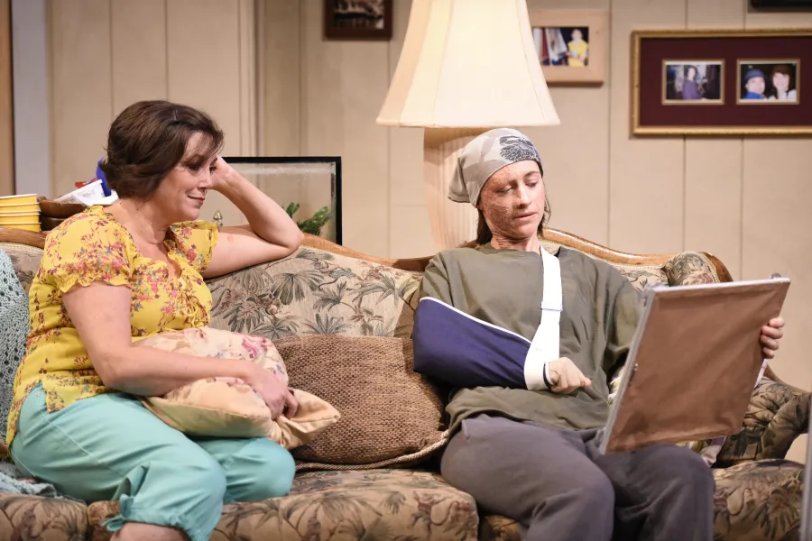 Two people sit on a couch; one wears a head wrap and arm sling while holding a framed photo, and the other, smiling, rests her arm on a pillow.