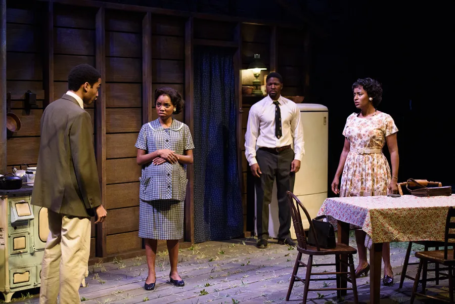 Four actors stand in a kitchen set on stage, engaged in conversation; kitchen appliances and a table are visible in the background.