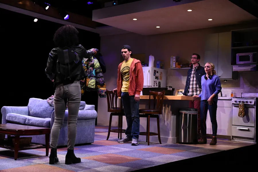 Four people are in a modern living room and kitchen set on stage; one person stands facing three others, who look toward her with varying expressions.