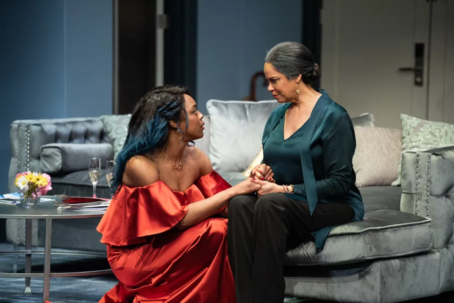 Two women sit on a sofa, holding hands and making eye contact; one wears a red dress, the other wears a dark green top. A table with flowers and drinks is beside them.