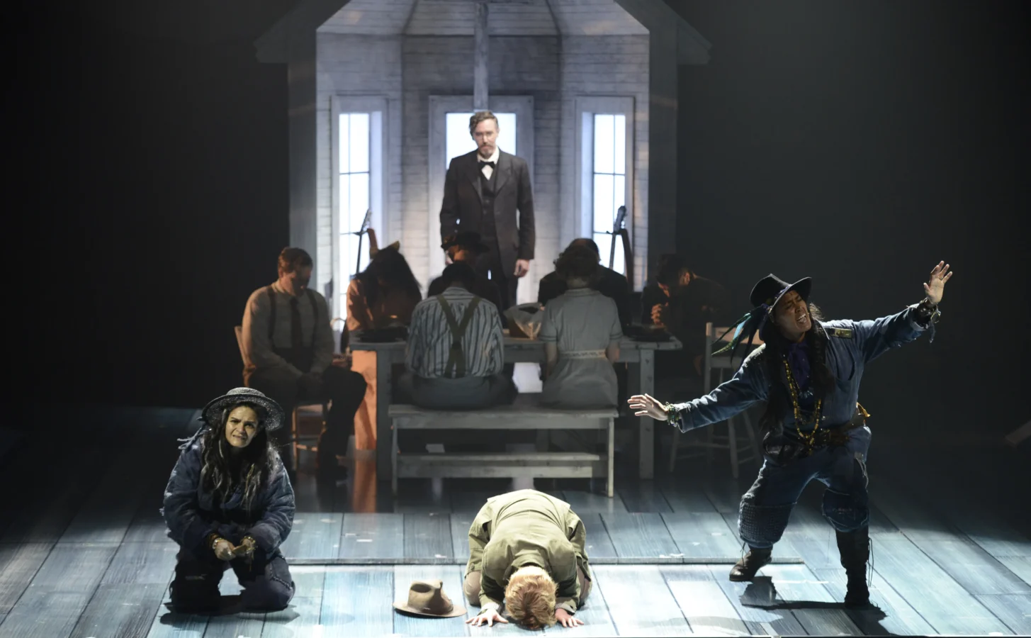 A theatrical performance shows actors on stage with a house backdrop; three performers are in dynamic poses at the front, while others sit at a table under dramatic lighting.