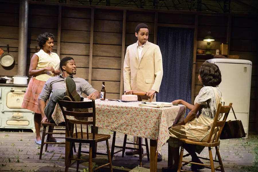 Four people interact around a table in a kitchen setting on stage, with a cake and bottle on the table and a vintage stove and refrigerator in the background.