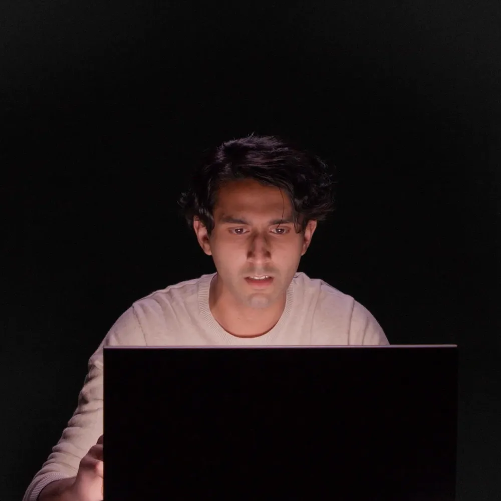 A person with dark hair looks intently at a laptop screen in a dark room, illuminated by the light from the screen.
