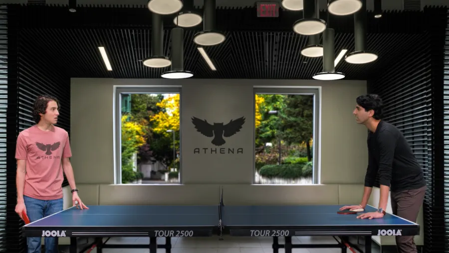 Two men stand on opposite sides of a ping pong table indoors, with the ATHENA logo displayed on the wall between two windows behind them.