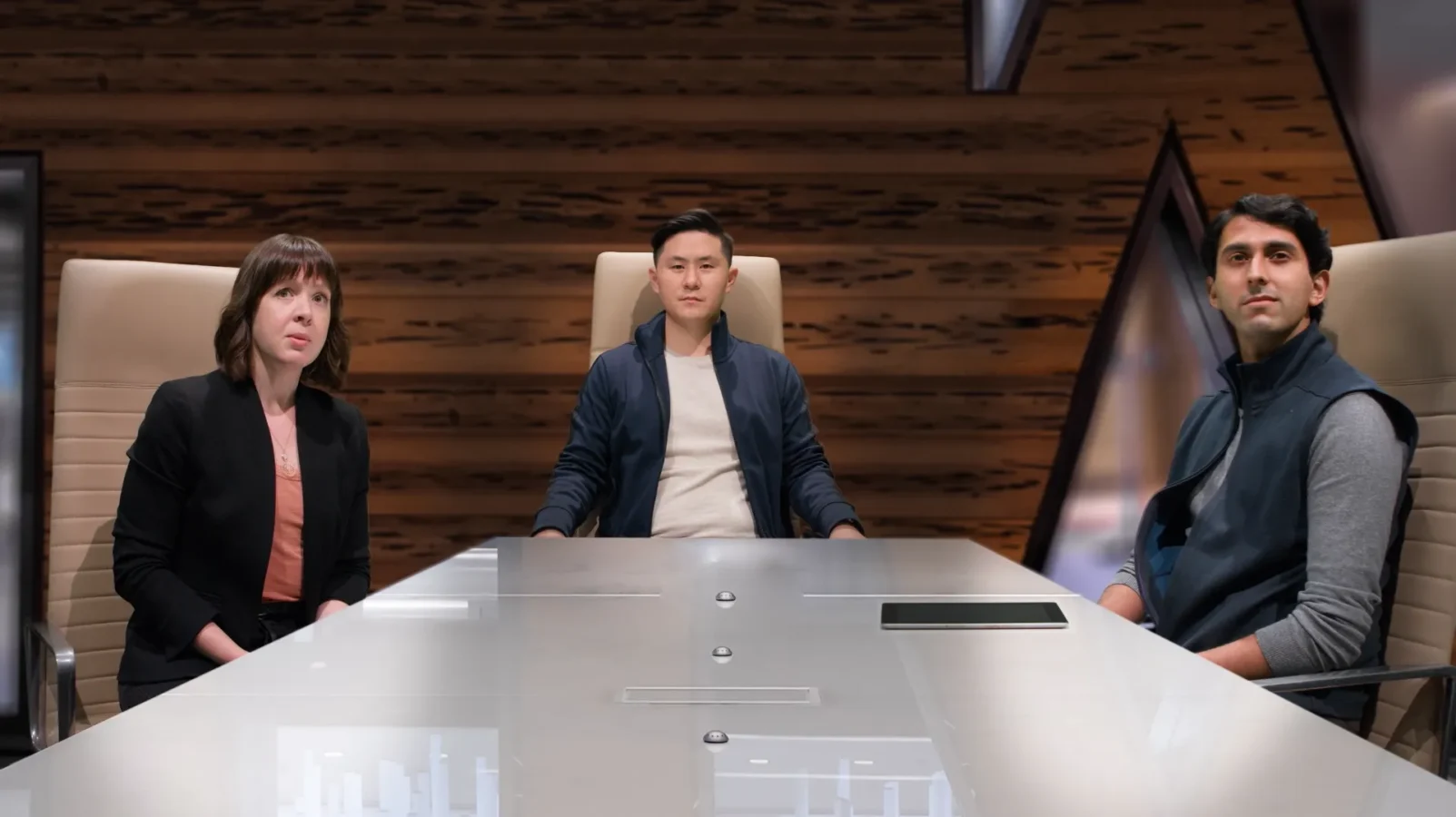 Three people sit at a conference table in a modern office, looking forward with neutral expressions.
