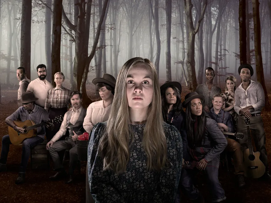 A group of people in period clothing pose in a foggy forest, with a young woman standing in the foreground and the rest sitting or standing behind her, some holding musical instruments.