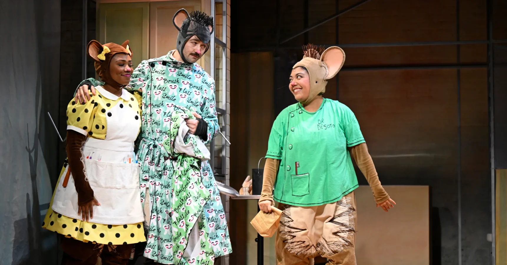 Three people dressed in colorful mouse costumes perform on stage with painted backgrounds and props, suggesting a theater production or play scene.
