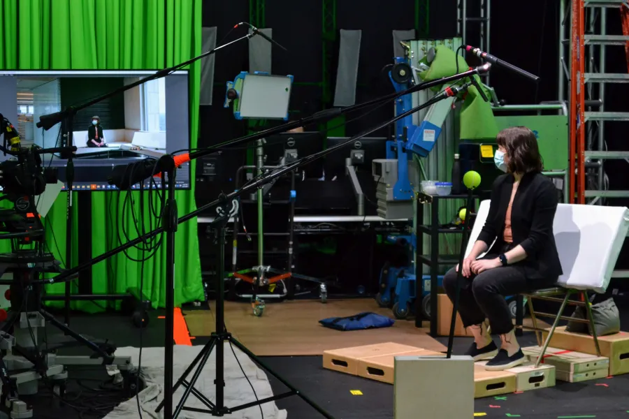 A person wearing a mask sits on a chair in a studio with green screens, cameras, microphones, and lighting equipment visible around them.