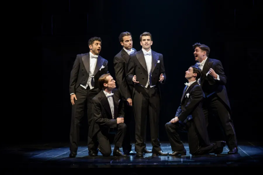 Six men in tuxedos perform on stage under dramatic lighting; four stand while two kneel, all engaged in expressive poses and facial expressions.