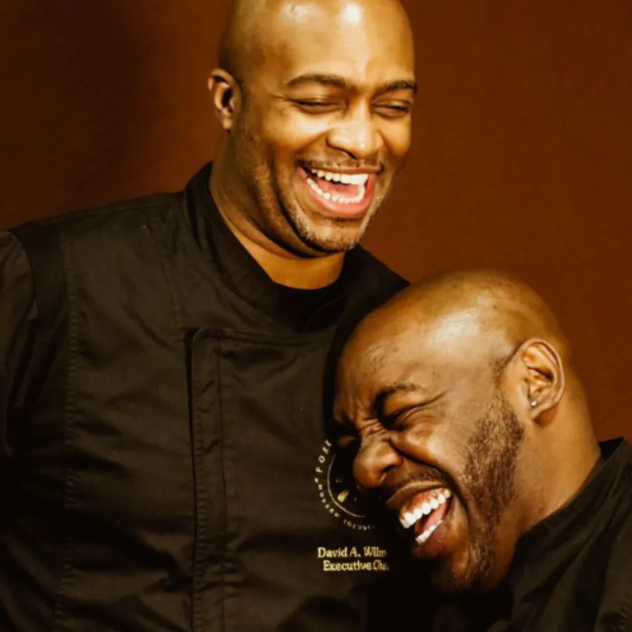 Two men wearing black chef coats laugh together, with one standing and the other leaning in close, both appearing joyful.