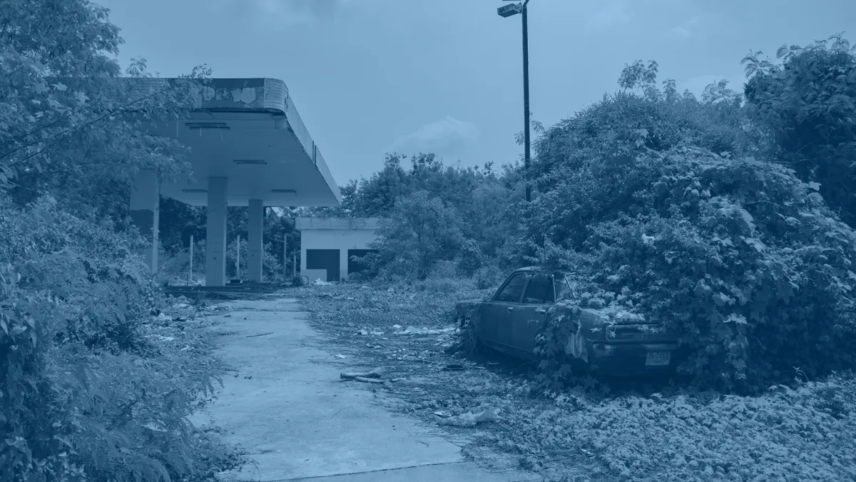 An abandoned gas station with overgrown vegetation, an old car partly covered by plants, and debris scattered on the ground under a cloudy sky.
