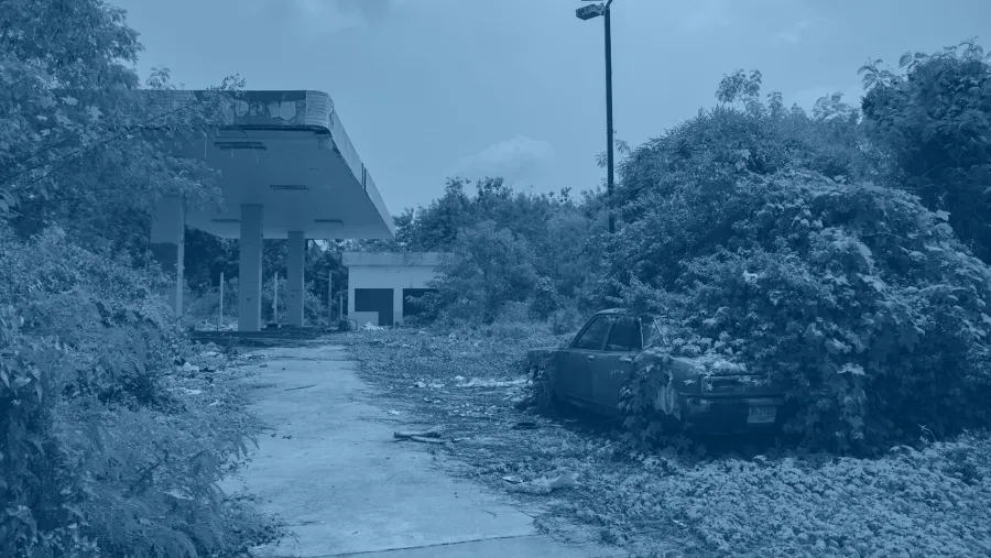 An abandoned gas station with overgrown vegetation and a rusted car partially covered by plants in the foreground.