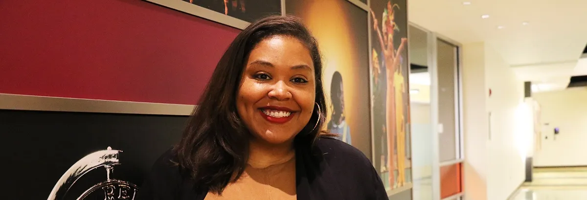A woman with long dark hair smiles while standing in a hallway, with framed posters and a maroon wall in the background.