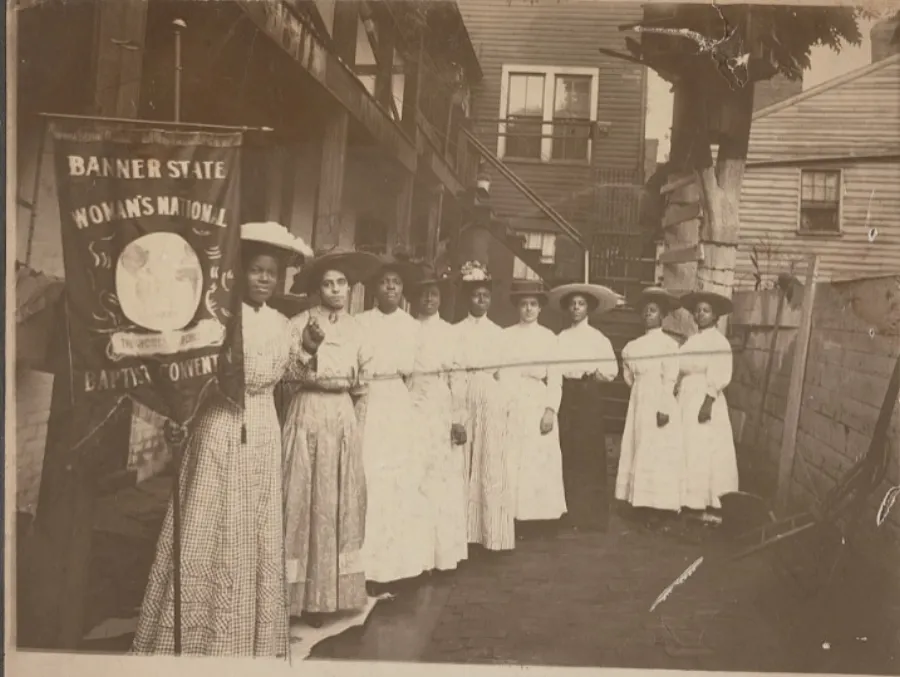 A group of women in dresses and hats stand in a row in an alleyway, holding a banner that reads "Banner State Woman's National Baptist Convention.