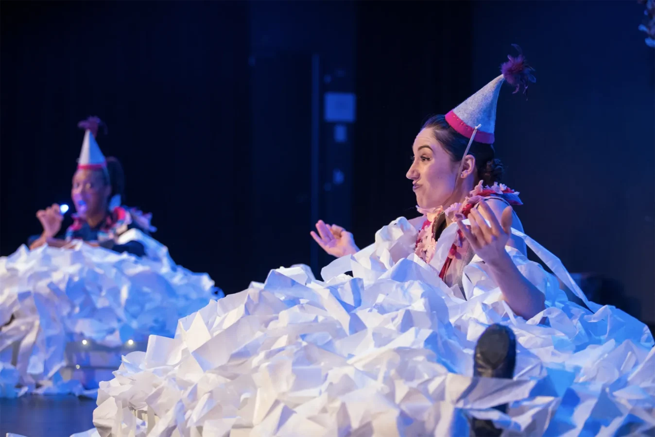 Two people wearing party hats sit amid piles of white paper strips on a stage, both making expressive gestures under blue stage lighting.