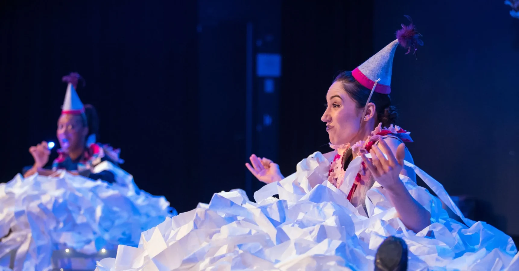 Two people wearing party hats and leis sit on stage, surrounded by piles of crumpled white paper, with one person gesturing expressively.