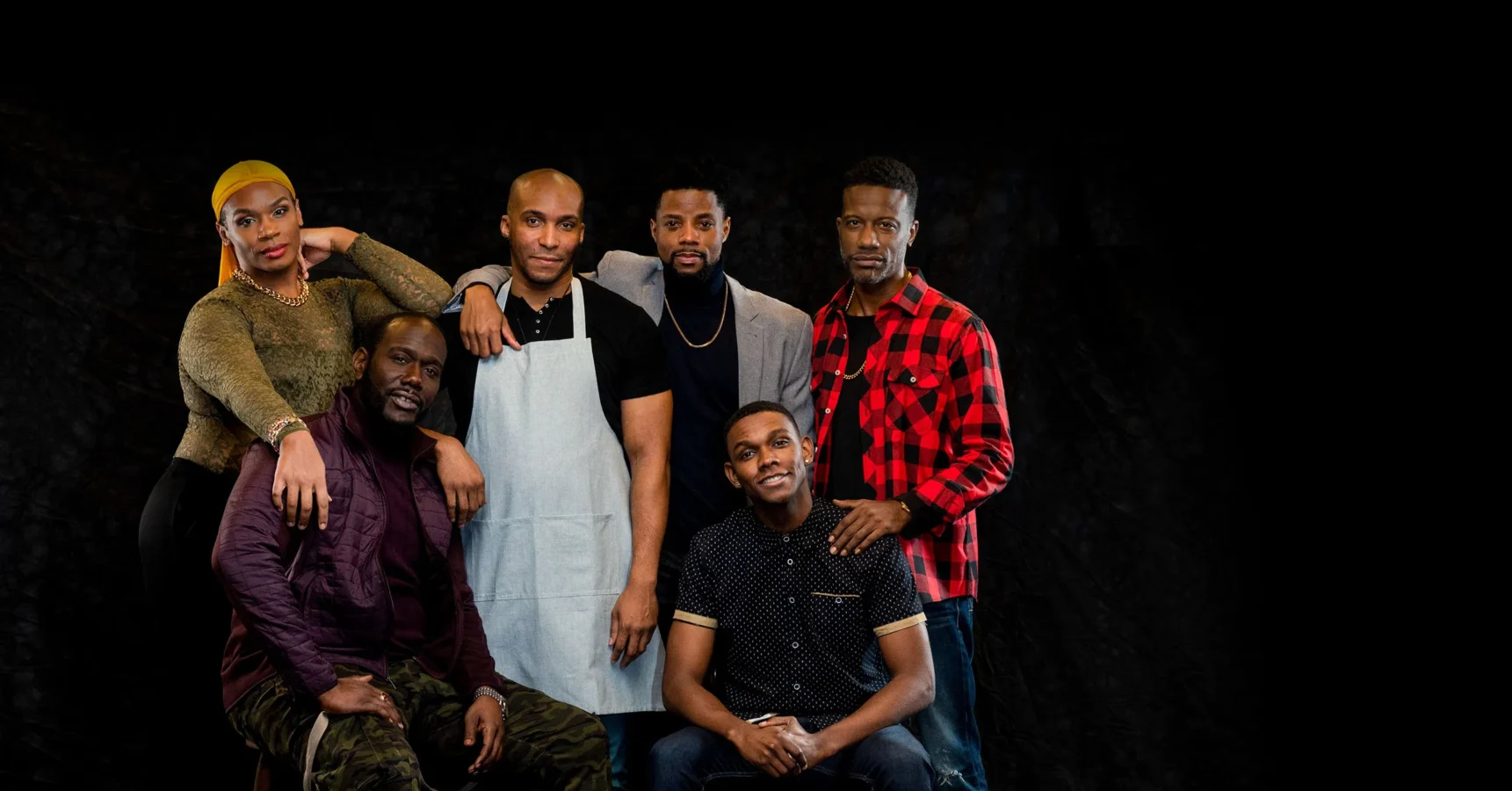 Six people pose together against a black background; one wears an apron, another sports a red plaid shirt, while the rest are dressed casually.