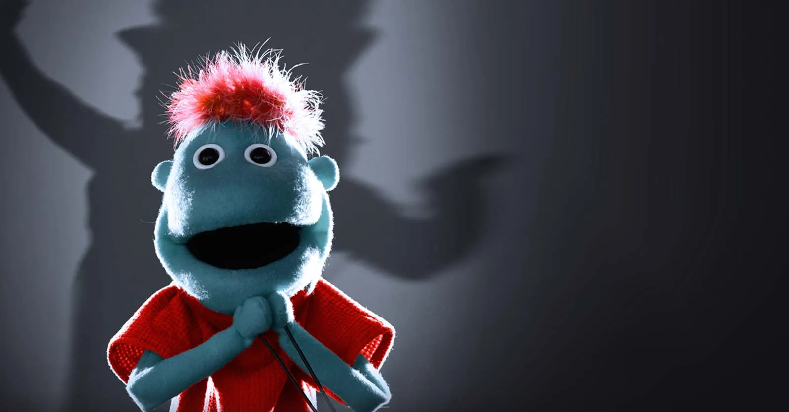 A blue puppet with red hair and a red sweater stands in front of a dark background, hands clasped, with its shadow cast on the wall behind it.
