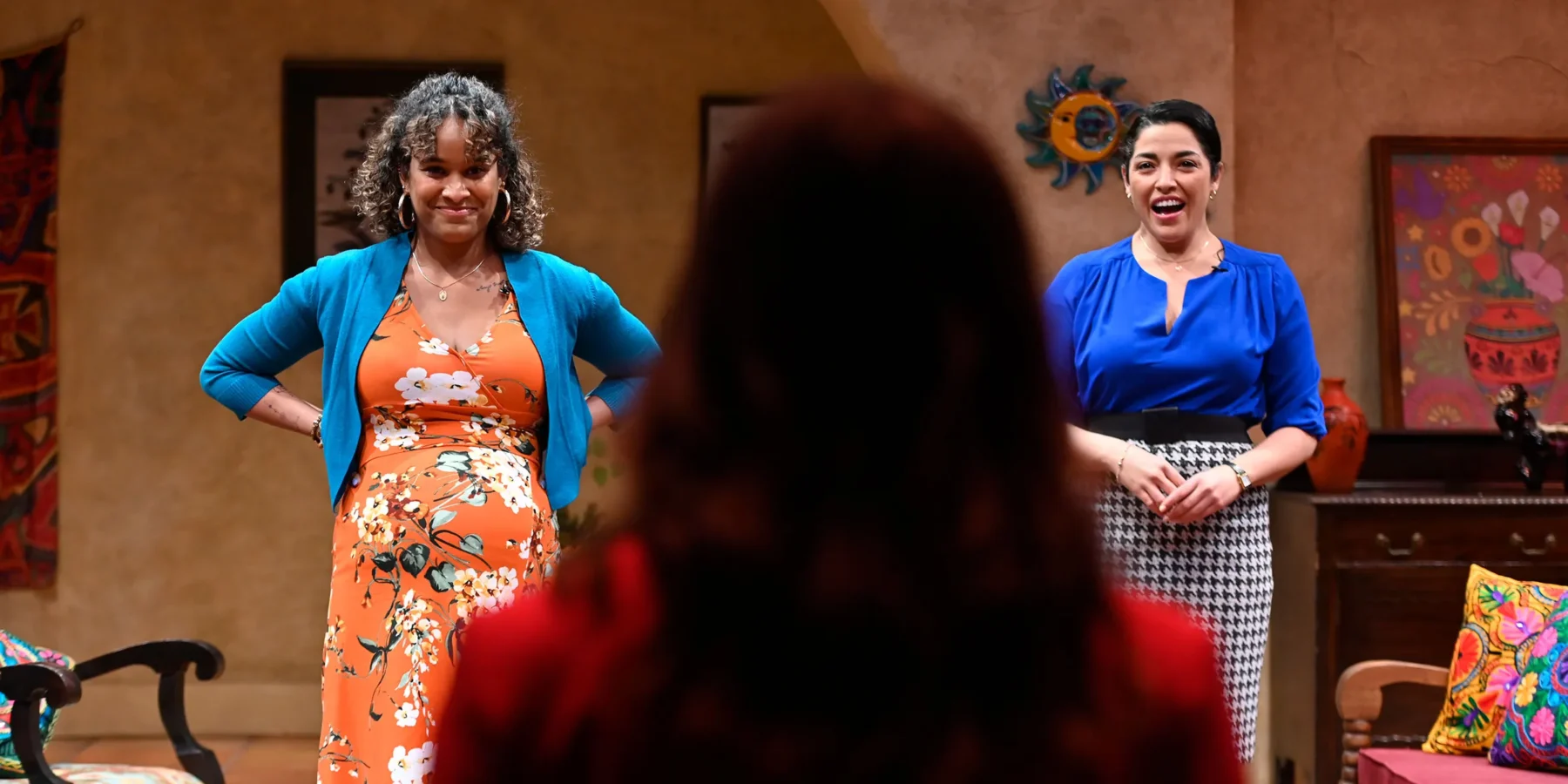 Two women stand facing a third person whose back is to the camera, in a brightly decorated room with colorful artwork and textiles.