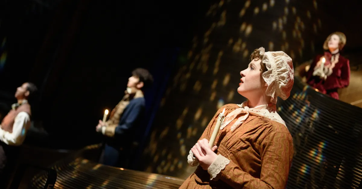 Four people in period costumes hold candles on stage, illuminated by warm, dappled lighting, suggesting a theatrical performance set in a historical era.