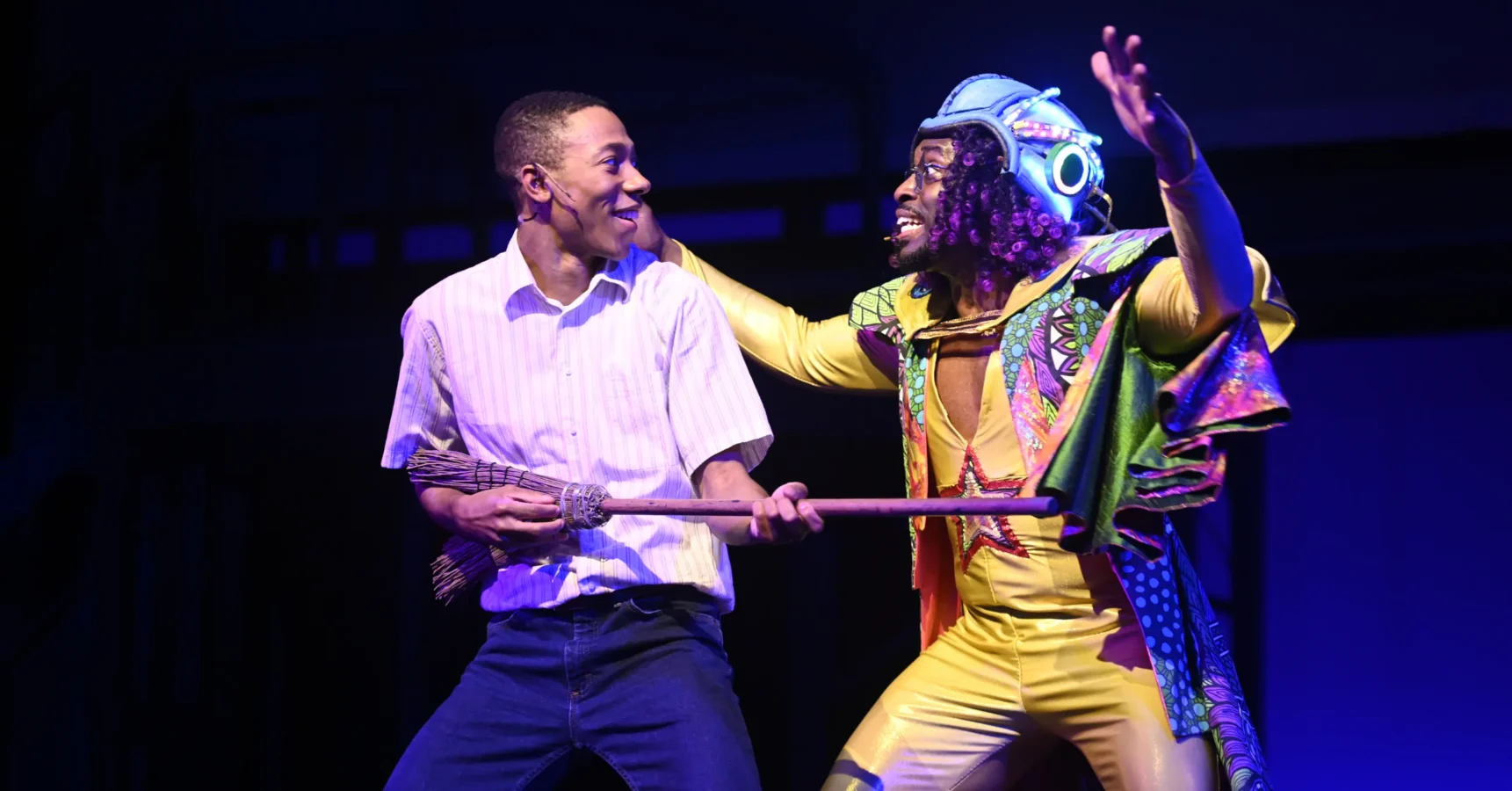 Two actors perform on stage; one holds a broom and wears casual clothes, while the other wears a colorful, futuristic costume and helmet.