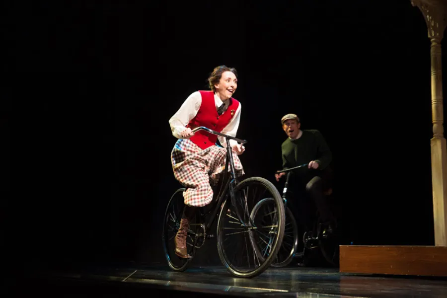Two actors on stage ride bicycles; the woman in front wears a red vest and plaid skirt, while the man behind wears a cap and dark sweater.