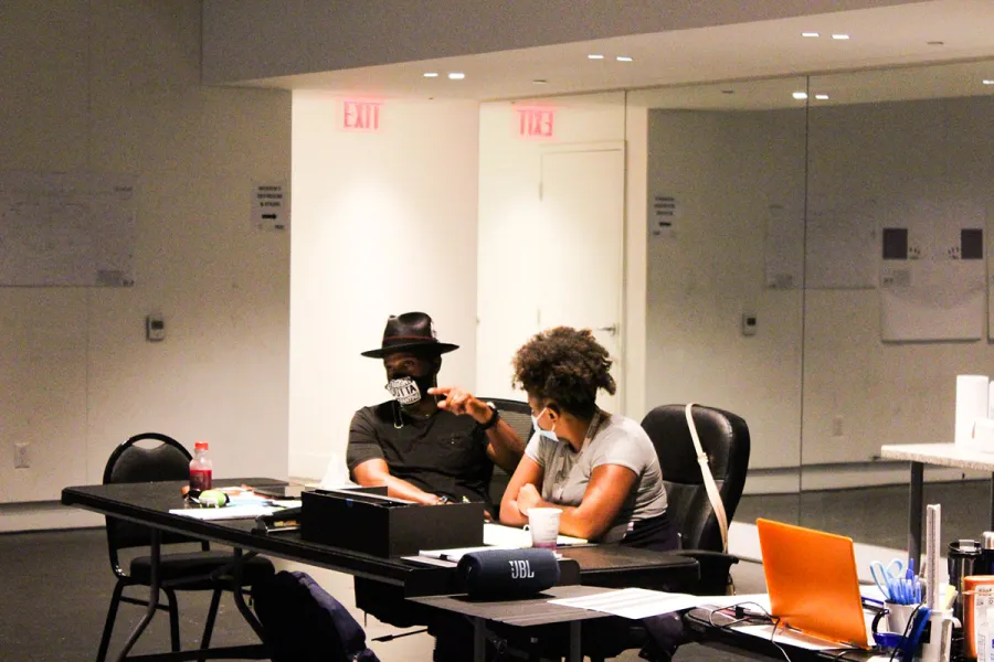 Two people sit at a table in an office or studio, engaged in conversation. Both are wearing face masks. The room has white walls and EXIT signs are visible in the background.