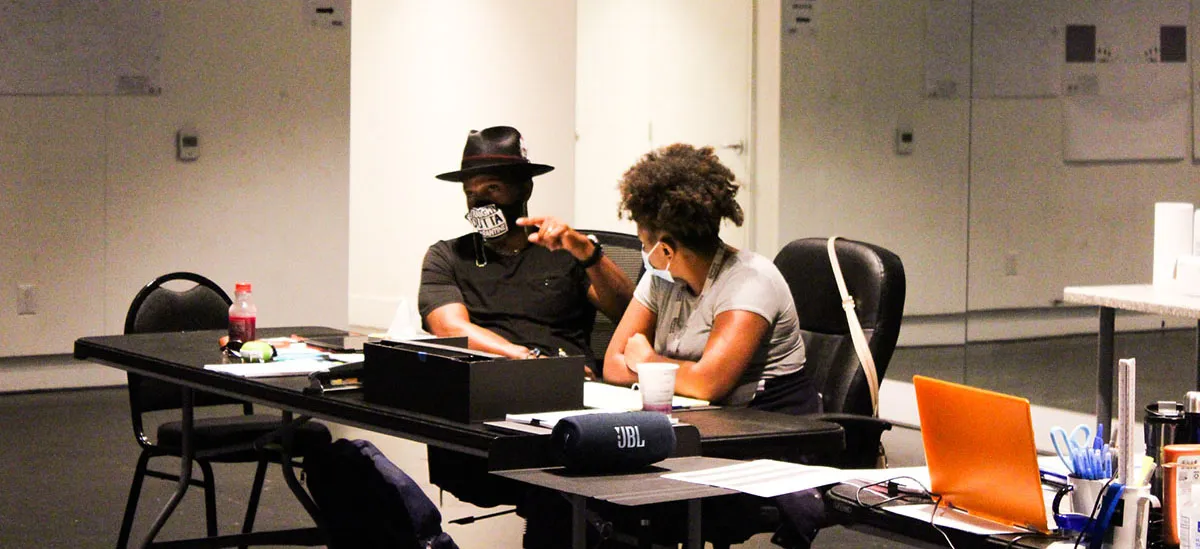 Two people sit at a desk in an office, conversing. One wears a hat and mask, the other has curly hair. Papers, a laptop, and office supplies are on the desk.