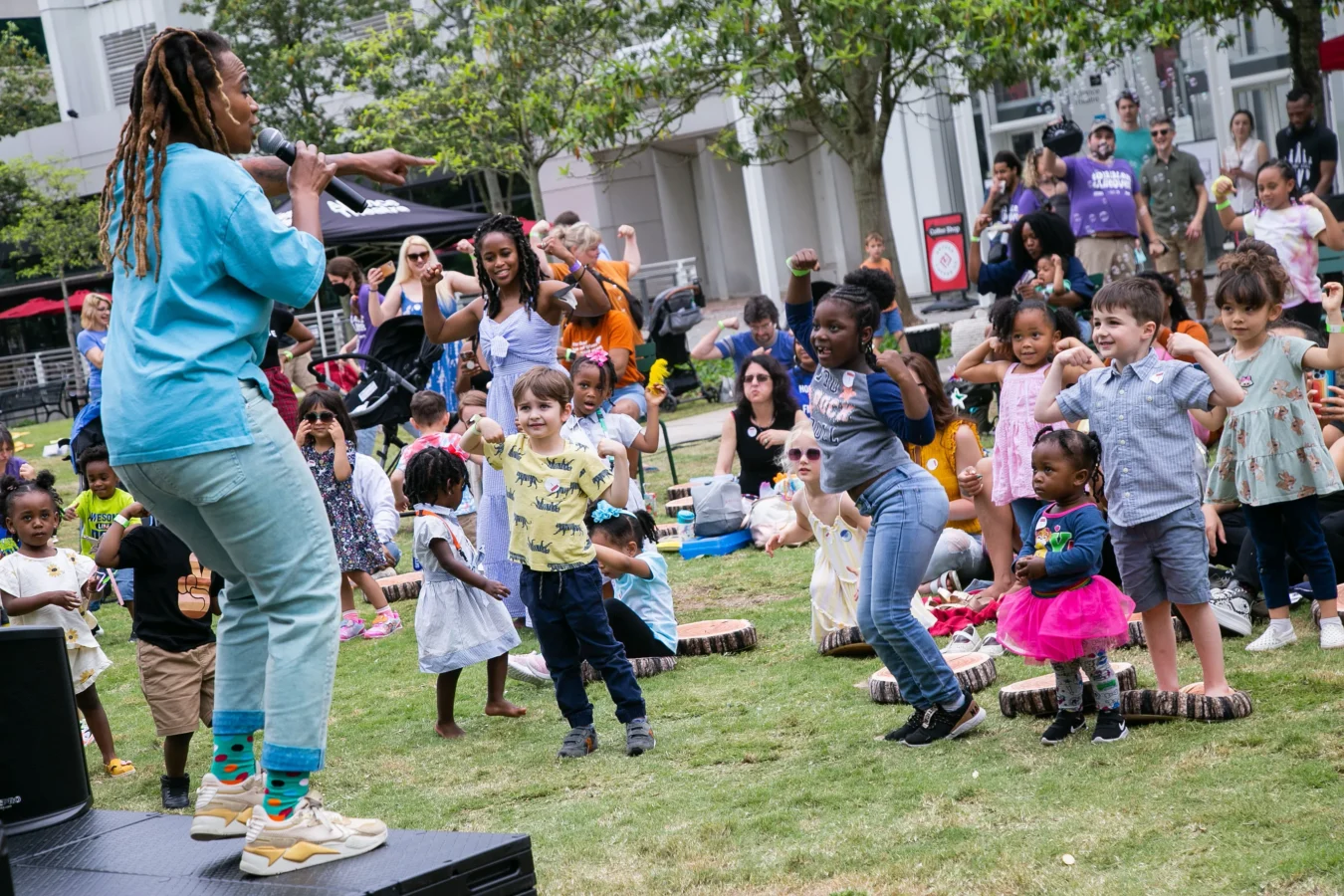 A performer with a microphone leads a group of young children in a dance activity outdoors, while adults supervise in the background.