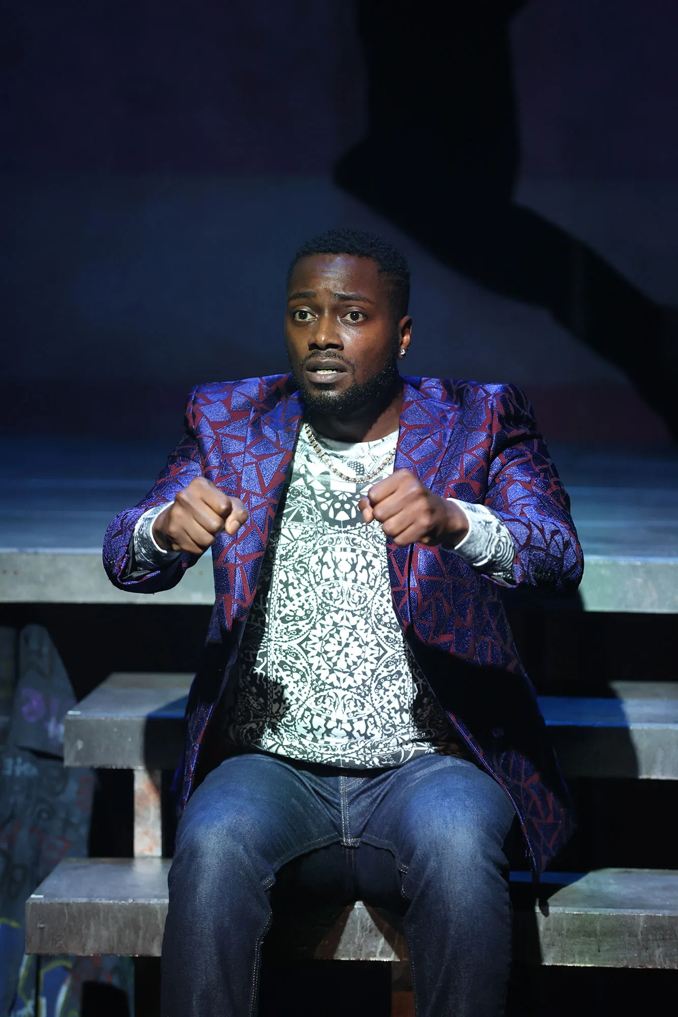 A man sits on steps, wearing a patterned blazer and jeans, holding his hands out as if gripping a steering wheel, with a concerned expression on his face.